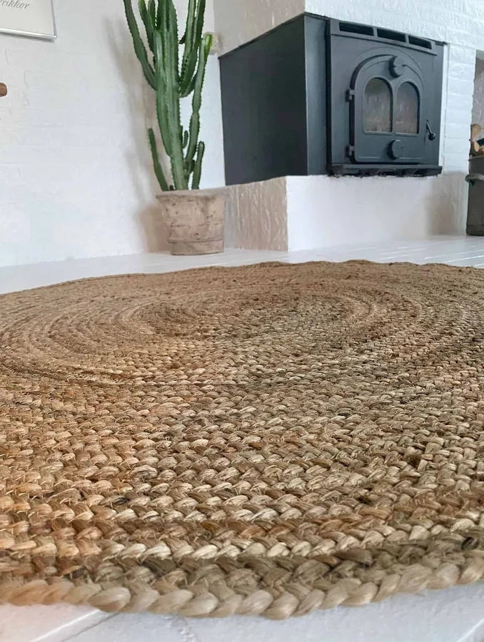 Jute vloerkleed rond naturel gevlochten 120 cm van de zijkant, op de achtergrond zie je een grote plant en een haard
