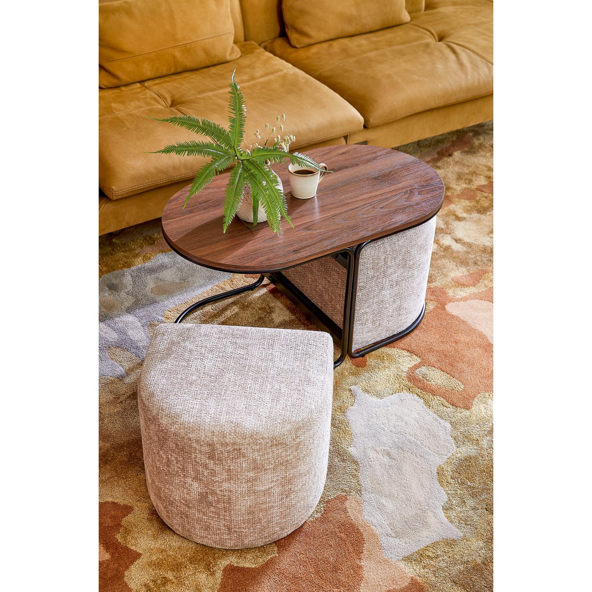 Closeup van een moderne salontafel met een ovale houten bovenkant en twee zachte beige poefs geïntegreerd in een zwart metalen frame. Het staat in een warm, sfeervol gelig interieur. De poefs kunnen los worden gebruikt als extra zitplaatsen of voetenbankjes. Een stijlvolle en multifunctionele toevoeging aan elk eigentijds interieur.