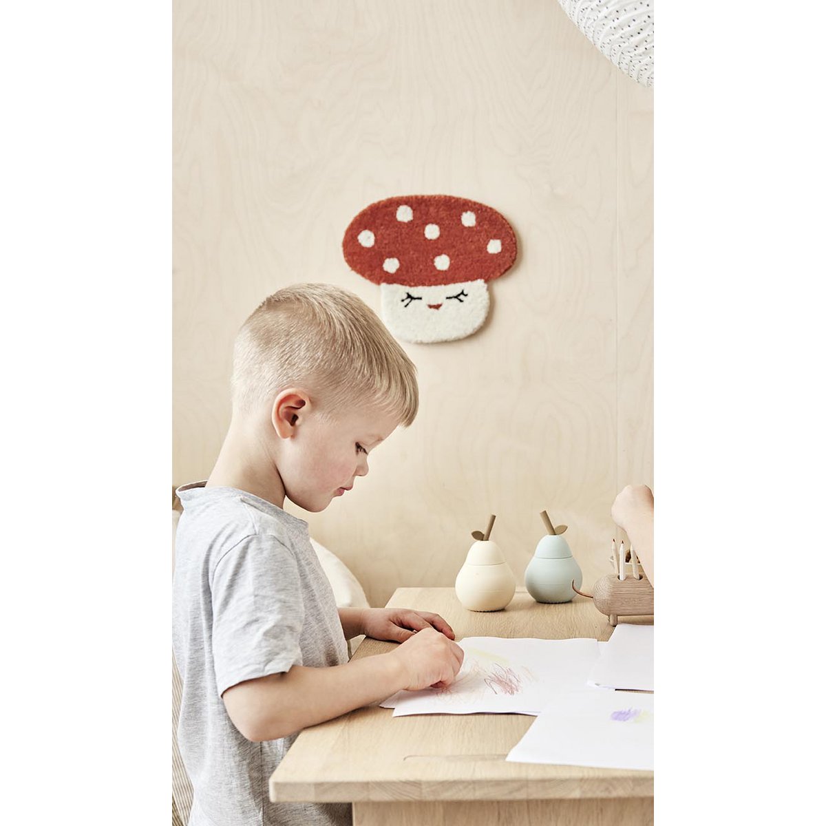 Een kind tekent aan een houten kindertafel met natuurlijke accessoires en een paddenstoel wanddecoratie in Scandinavische stijl.