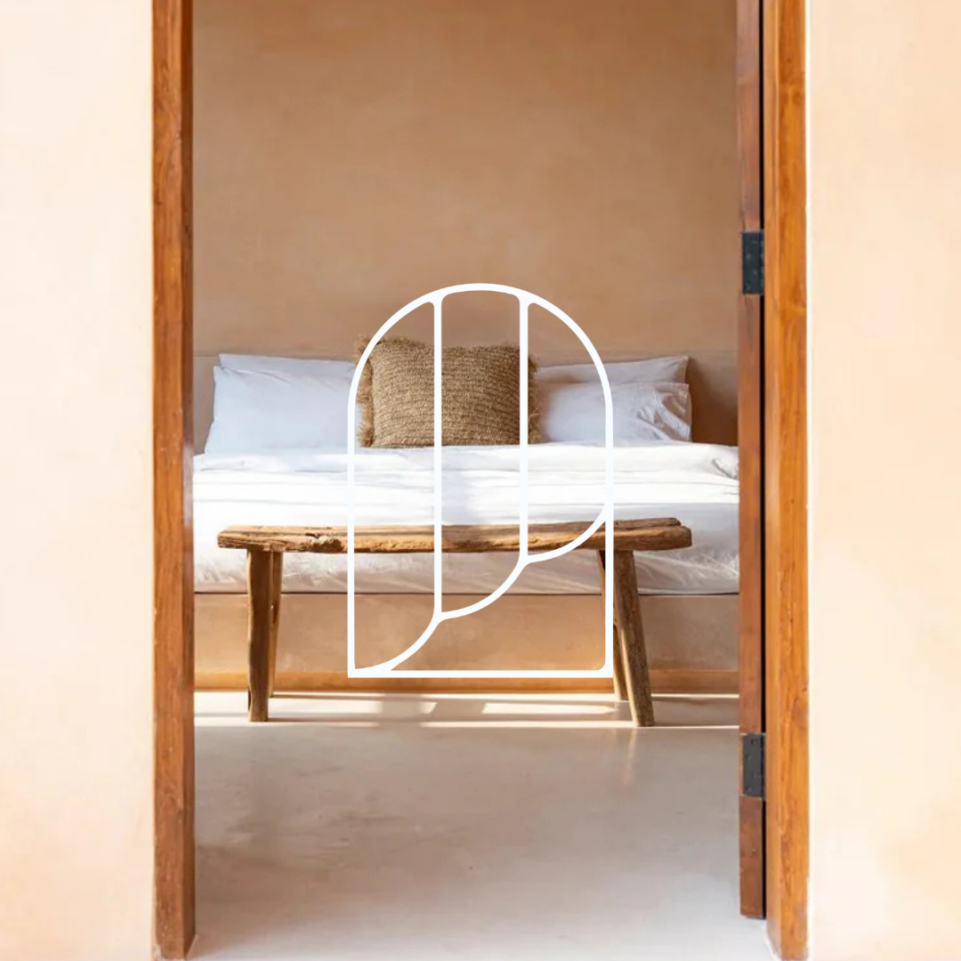 Afbeelding uit de collectie Plaids & Dekentjes. Op de foto zie je een deurframe met daarachter een slaapkamer. Daar zie je een bed met witte en beige kussens, en een witte deken erover. Daarvoor zie je een houten krukje.