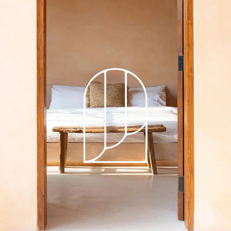 Afbeelding uit de collectie Plaids & Dekentjes. Op de foto zie je een deurframe met daarachter een slaapkamer. Daar zie je een bed met witte en beige kussens, en een witte deken erover. Daarvoor zie je een houten krukje.