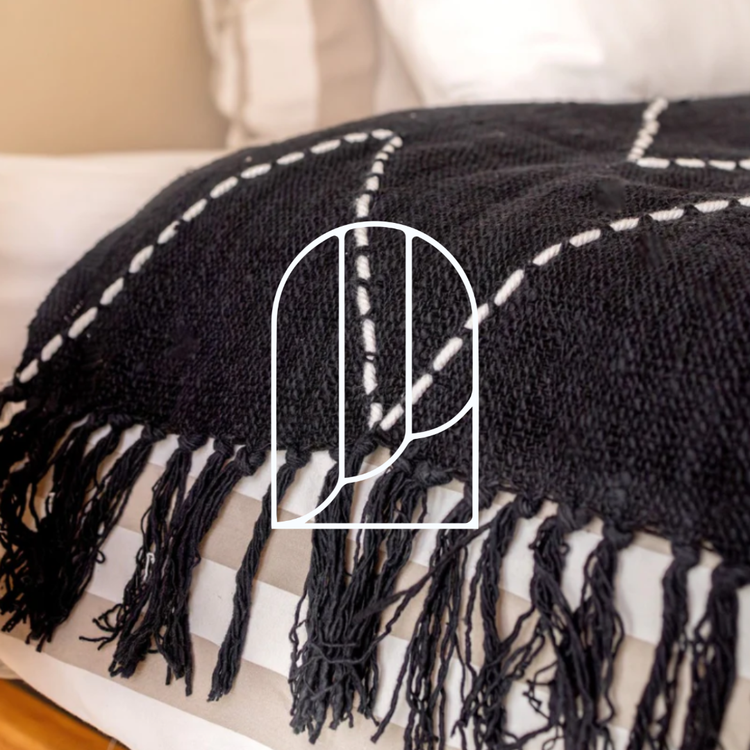 Afbeelding voor de collectie Dekens, Plaids & Bedspreien. Op de foto zie je een zwarte plaid met fransjes op het uiteinde van een bed.