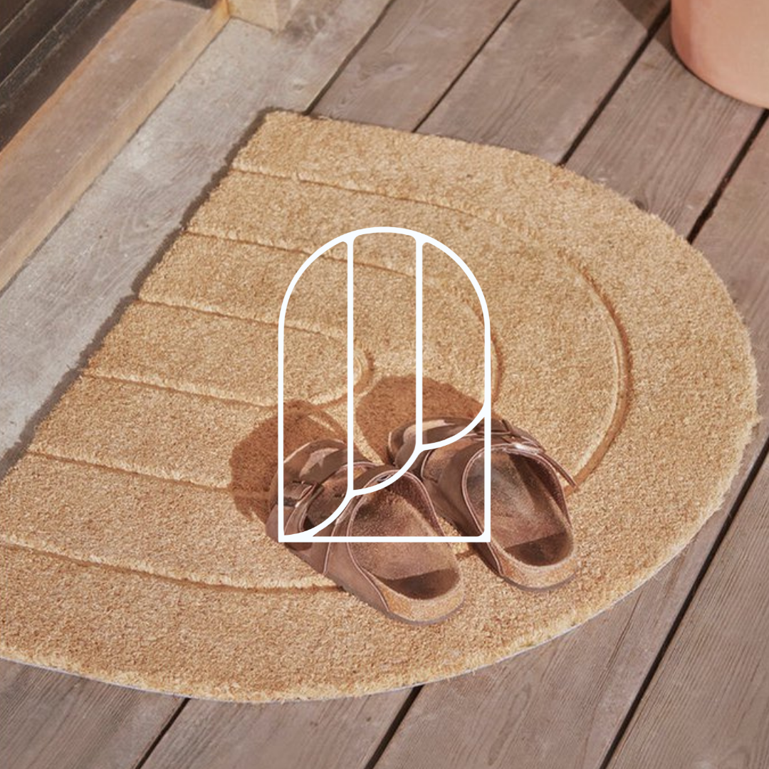 Afbeelding uit de collectie Deurmatten. Op de foto zie je een beige deurmat op houten vloer, met daarop een paar bruine slippers.