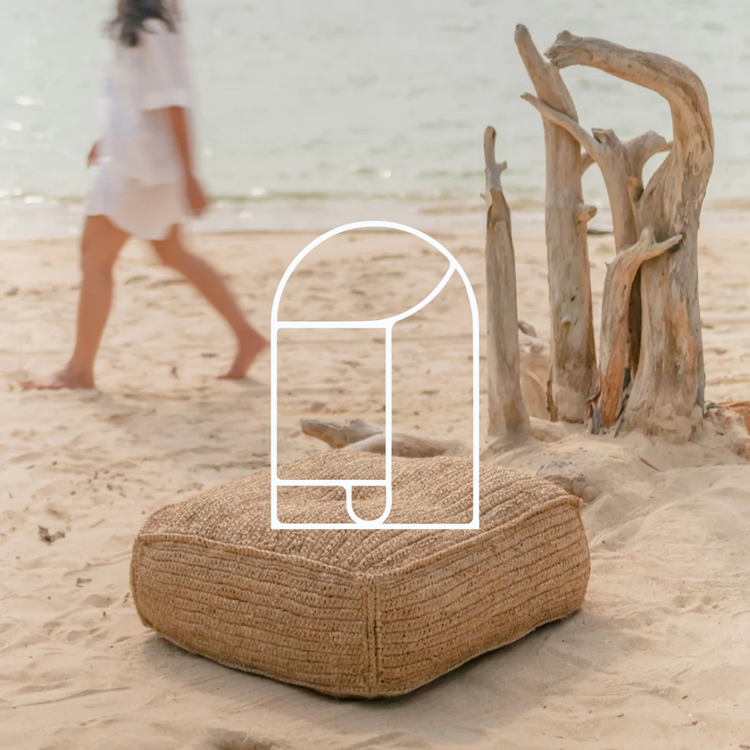 Afbeelding voor de collectie Poefs & Krukjes. Op de foto zie je een beige poef op het strand, met op de achtergrond boomresten, een vrouw die langs loopt en de zee.