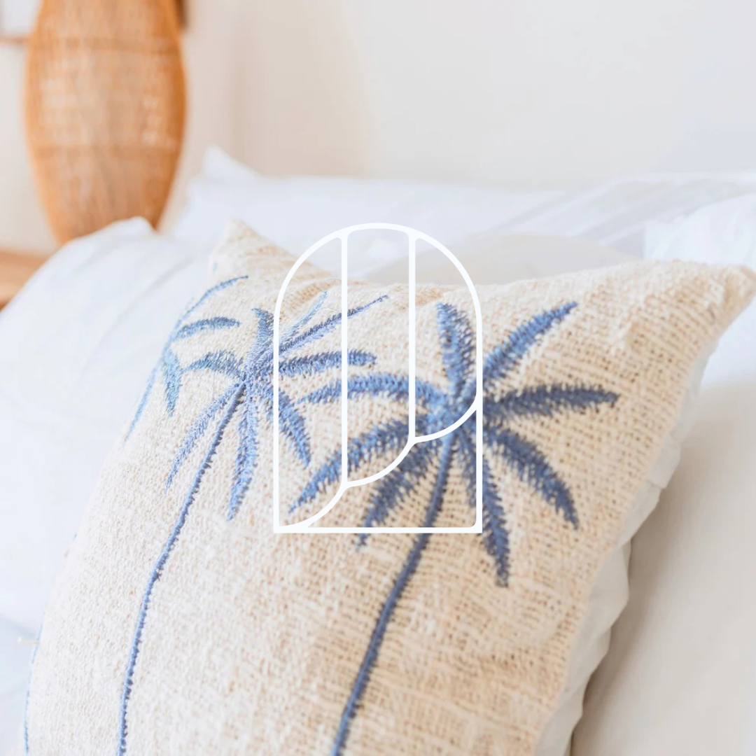 Afbeelding uit de collectie Sierkussens & Binnenkussens. Op de foto zie je een wit kussen met blauwe palmbomen erop op een wit opgemaakt bed.