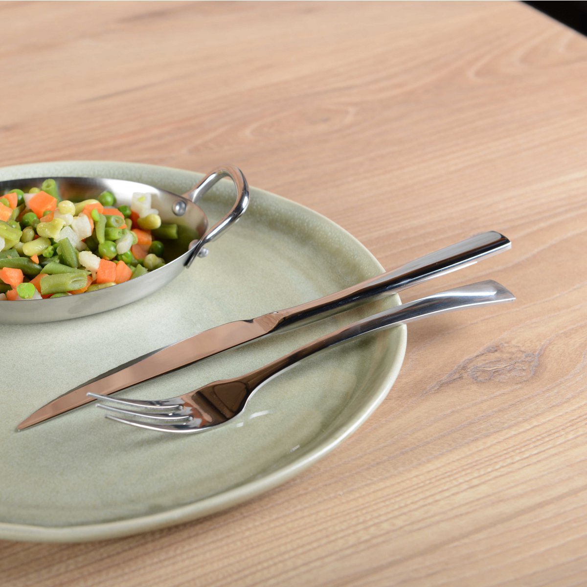 Bestekset zilver roestvrij staal 12-delig 6 personen, hier een vork en mes op een groen bord met schaaltje met groenten