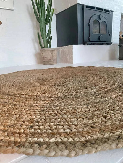 Jute vloerkleed rond naturel gevlochten 120 cm van de zijkant, op de achtergrond zie je een grote plant en een haard