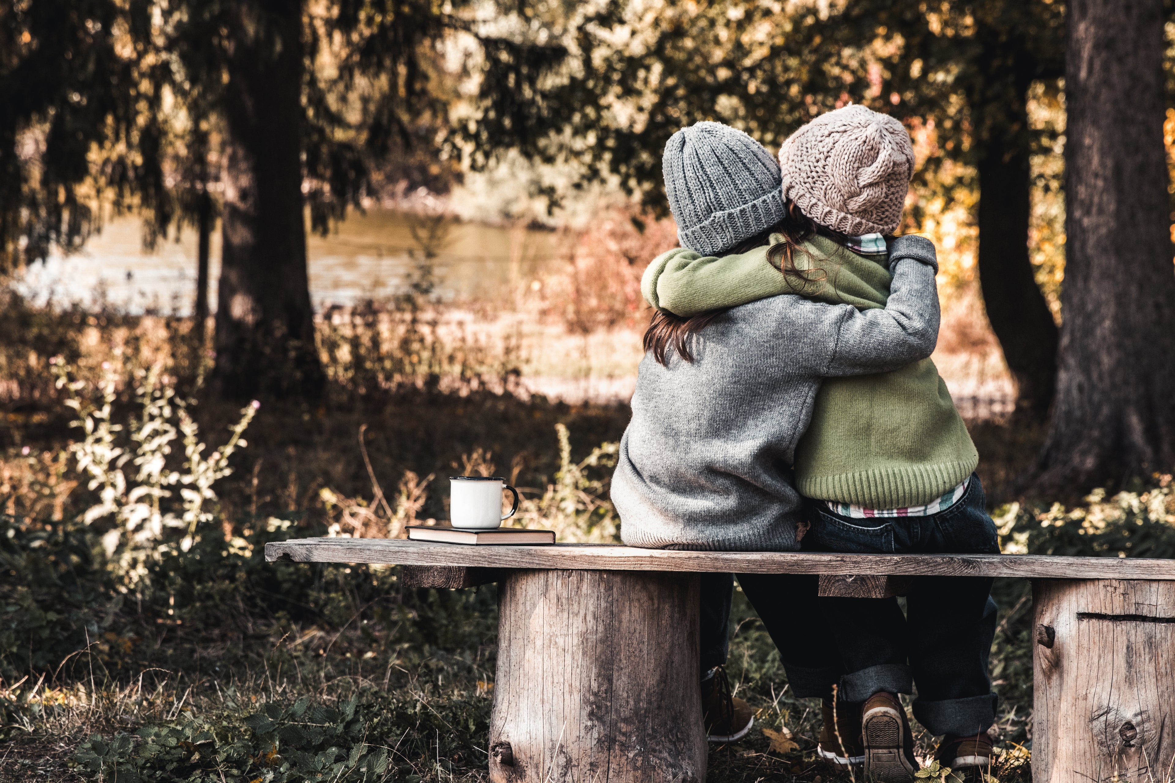 Twee mensen in warme truien en mutsen omarmen elkaar op een houten bankje in het bos, een moment van rust, verbondenheid en herfstinspiratie vol knus wonen, natuurlijke materialen en najaarssfeer van Luben.