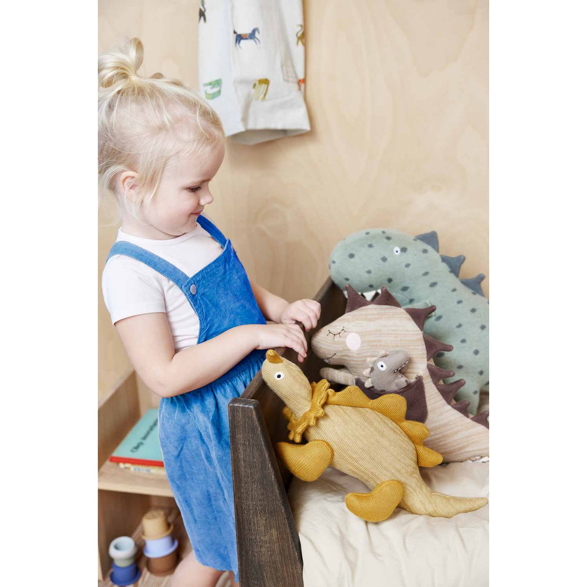 Knuffel dino Billy bruin in een kinderbed met een boel andere knuffels. Een meisje lacht naar de knuffels