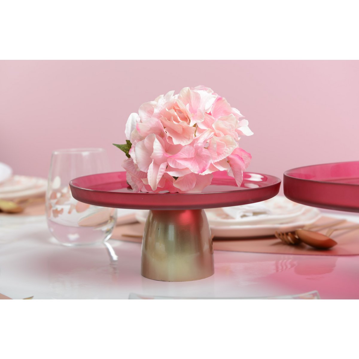 Rozerode glazen taartplateau, het standaard is goud. Omringt door wit en glazen servies, op de schaal ligt een mooie grote roze bloem ter decoratie 