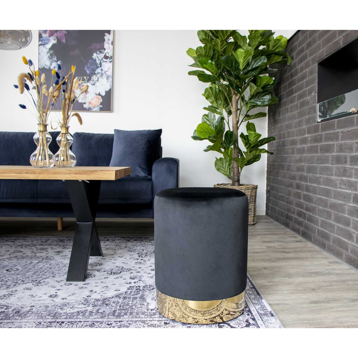 Zwarte fluwelen poef met goudkleurige basis in een moderne woonkamer met donkerblauwe bank, houten salontafel en groene kamerplant, een stijlvol woonaccessoire dat luxe en elegantie toevoegt aan het interieur.