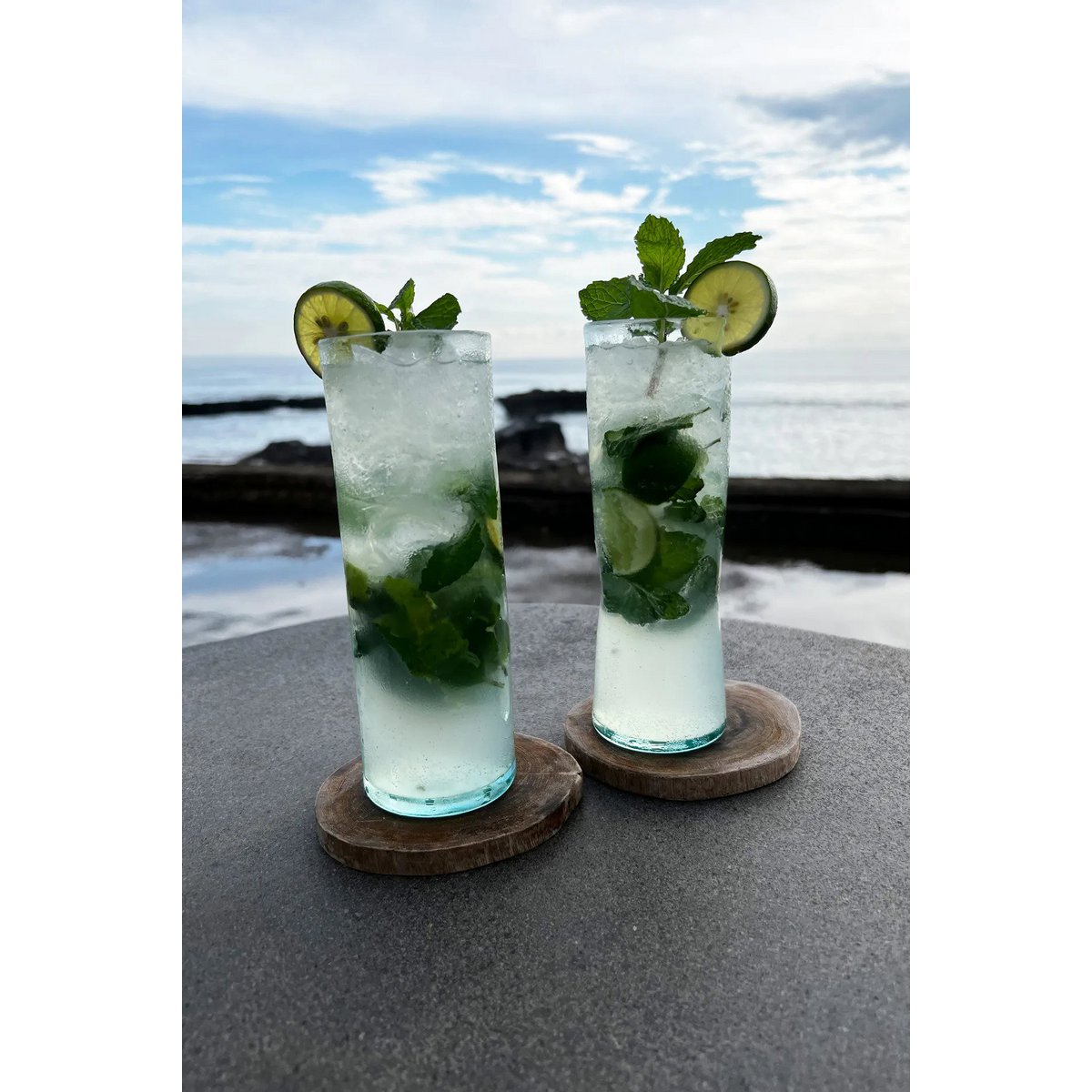 Twee teakhouten onderzetters met daarop twee glazen met ijskoude drankjes met op de achtergrond de zee