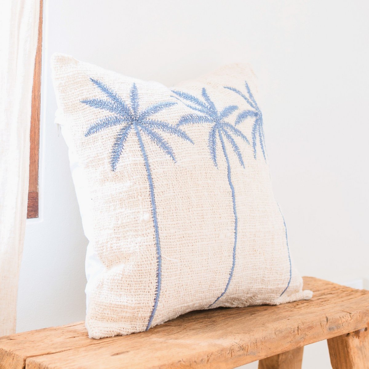 Witte kussenhoes met blauw palmmotief Anarka zijaanzicht op een houten kruk