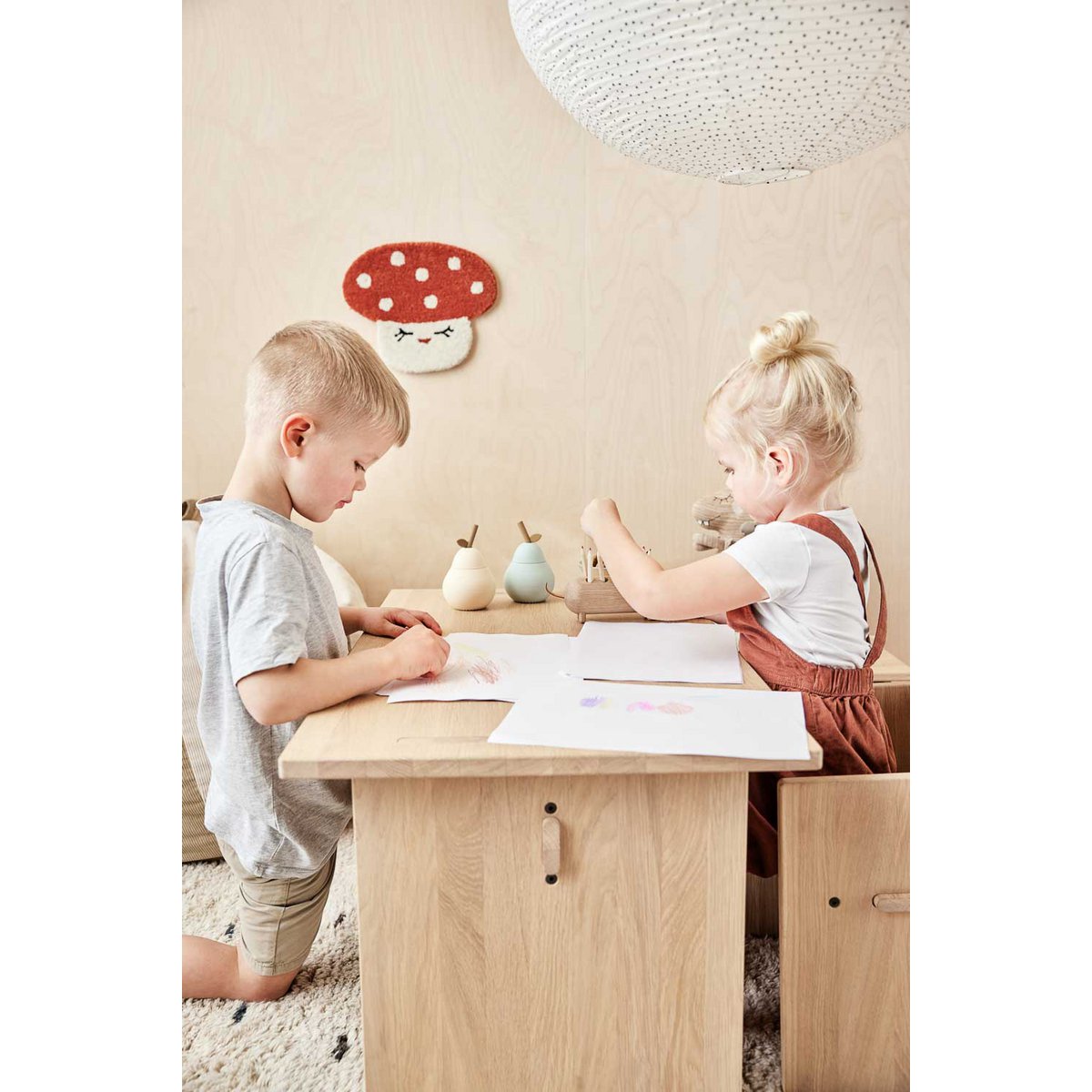 Twee kinderen tekenen aan een houten kindertafel met natuurlijke accessoires en een paddenstoel wanddecoratie in Scandinavische stijl.