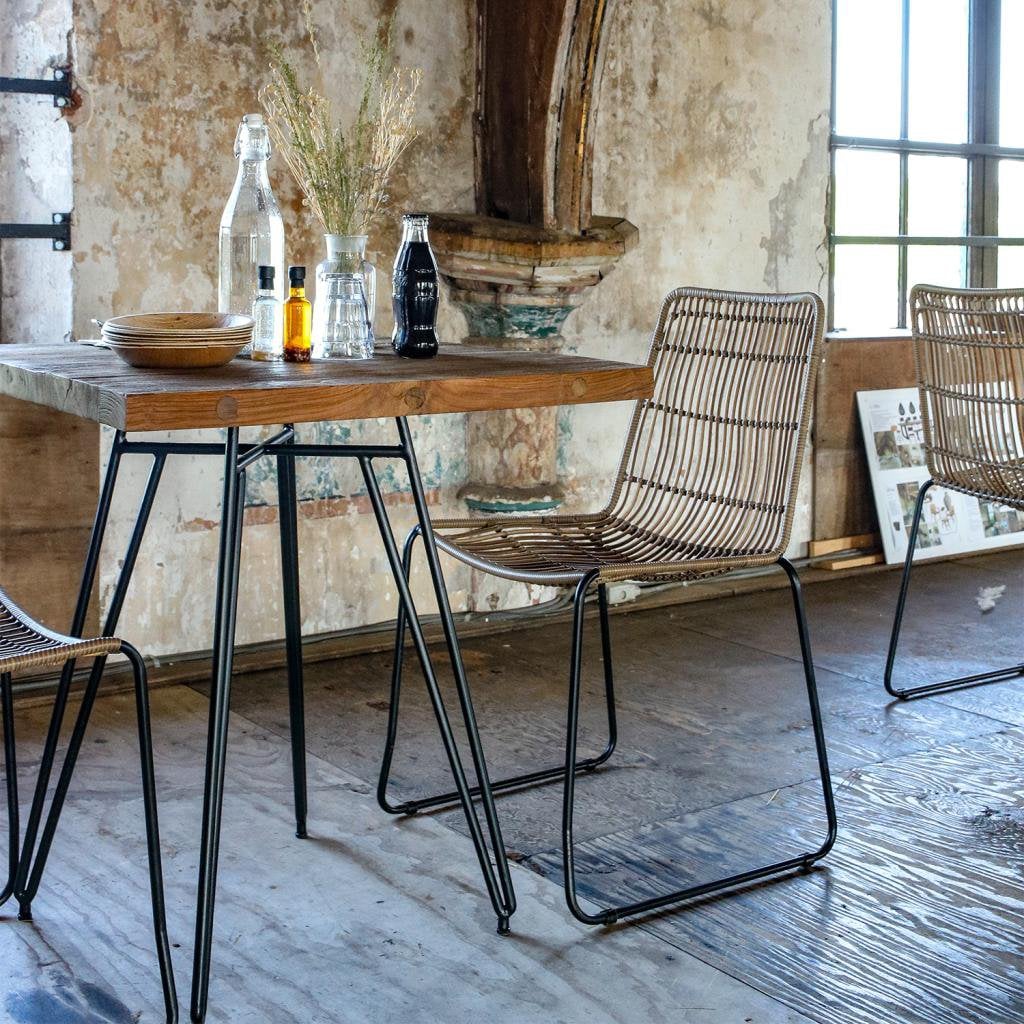 de Jane tuinstoel staat naast een houten tafel met zwart metalen frame, in een industriële ruimte met verweerde muren. Op de achtergrond nog een tweede stoel.