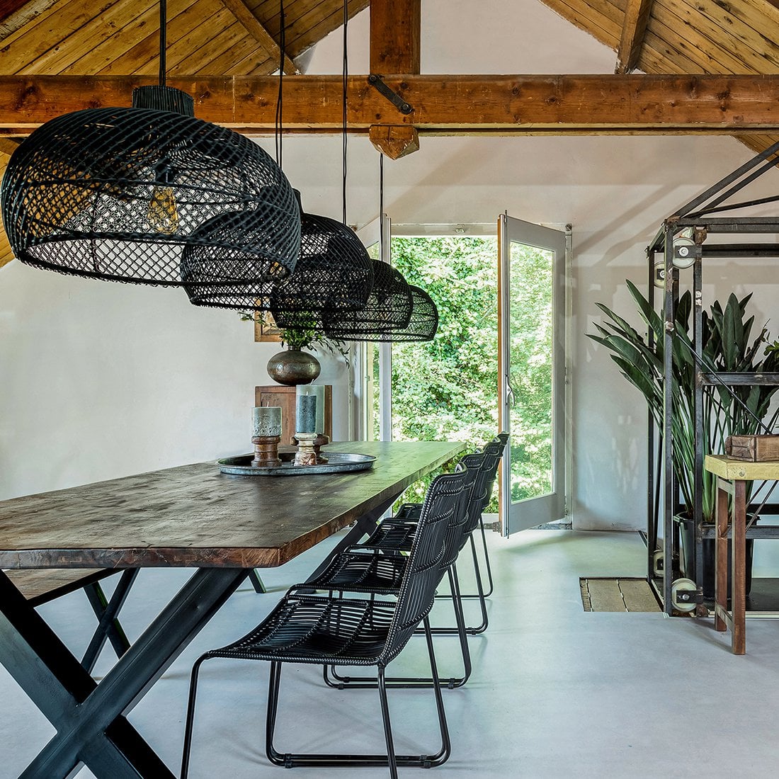 Meerdere zwarte rotan Jane stoelen aan een lange houten eettafel, met zwarte rotan hanglampen erboven in een lichte ruimte met houten dakbalken.