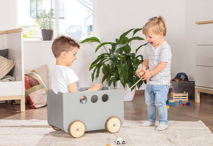 Kinderen spelen met grijze speelgoedkist op wielen – houten opbergmeubel en rijdend speelgoed in één.